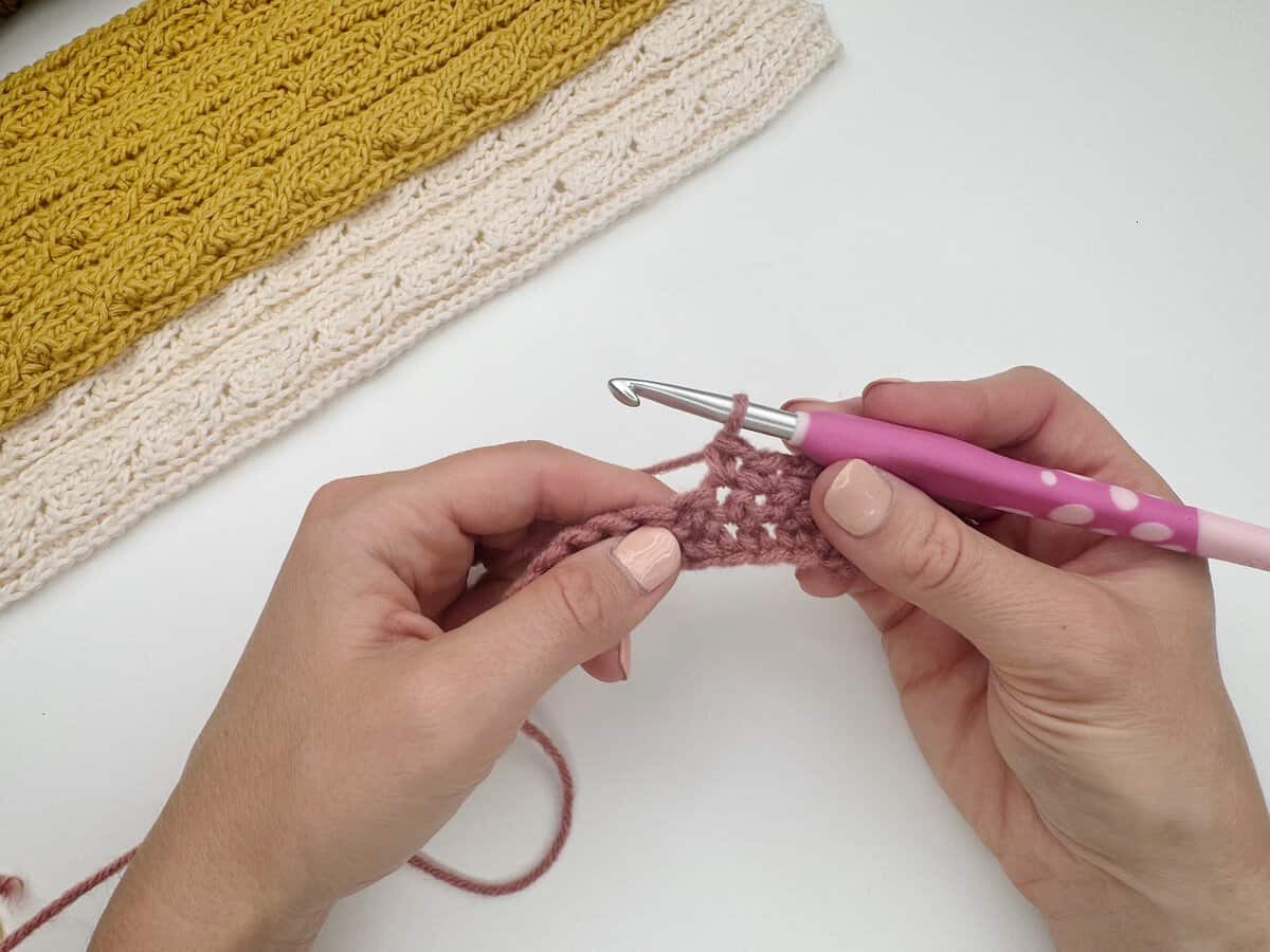 Hands holding a pink crochet hook and working with pink yarn, demonstrating a single crochet stitch tutorial; finished knitted pieces in yellow and cream sit in the background on a white surface.