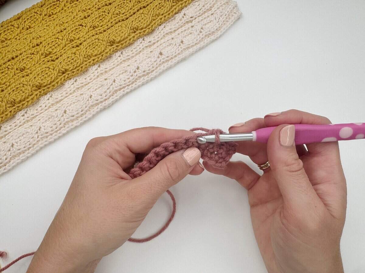 Close-up of hands demonstrating a single crochet stitch tutorial with pink yarn and a pink hook; finished sections of yellow and cream crochet work are also visible on the table.