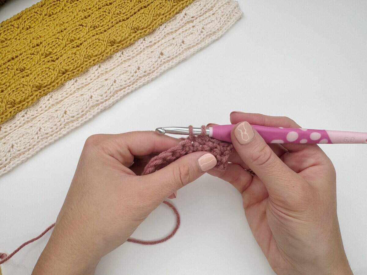 Close-up of hands crocheting with a pink hook and mauve yarn, demonstrating a single crochet stitch tutorial. Finished yellow and cream crochet pieces are visible in the background on a white surface.