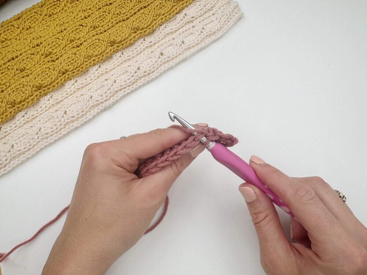 Close-up of hands crocheting with a pink-handled crochet hook and pink yarn, demonstrating a single crochet stitch tutorial, with a textured yellow and cream crocheted piece in the background.