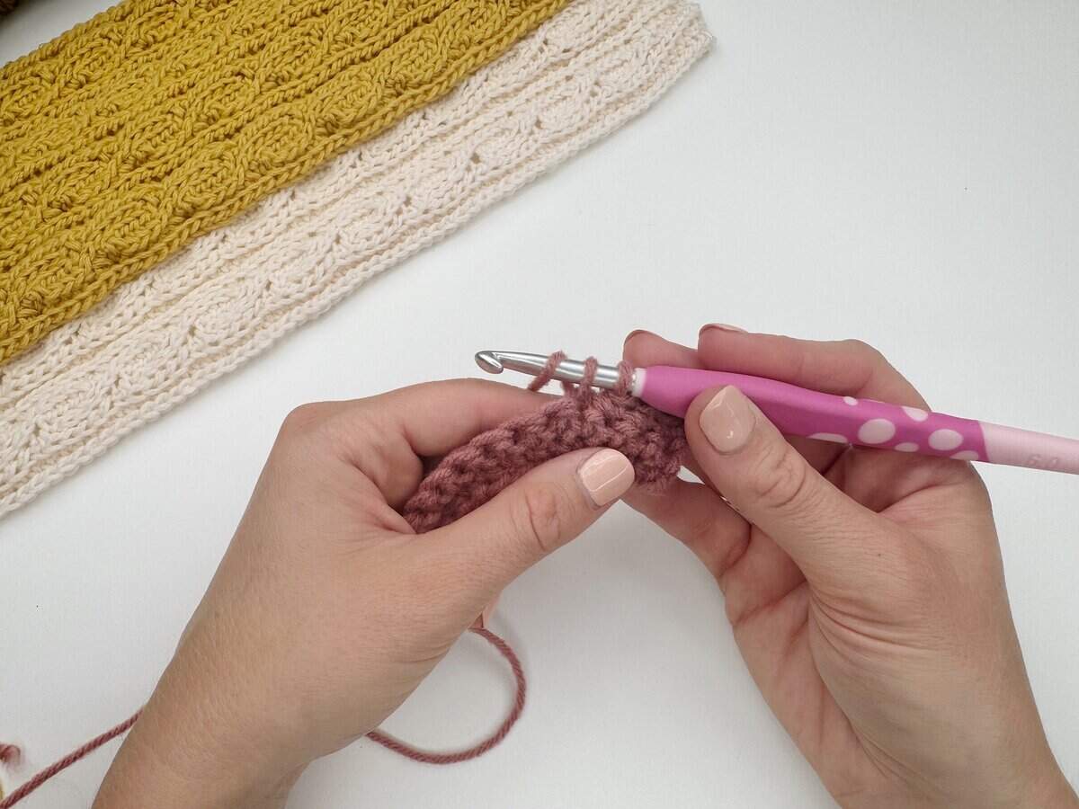 Two hands holding a crochet hook with pink yarn, following a single crochet stitch tutorial. Finished pieces in yellow and cream are visible in the background on a white surface.