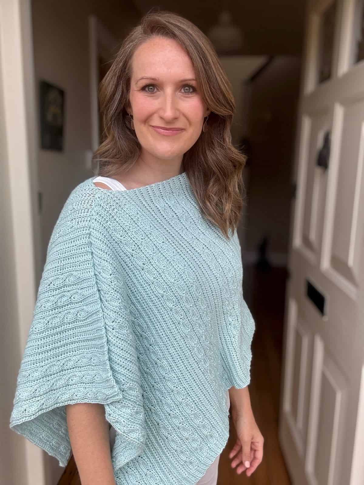 A woman with wavy brown hair stands in a doorway, wearing a light blue textured crochet poncho made from a free crochet poncho pattern, and smiles slightly at the camera.