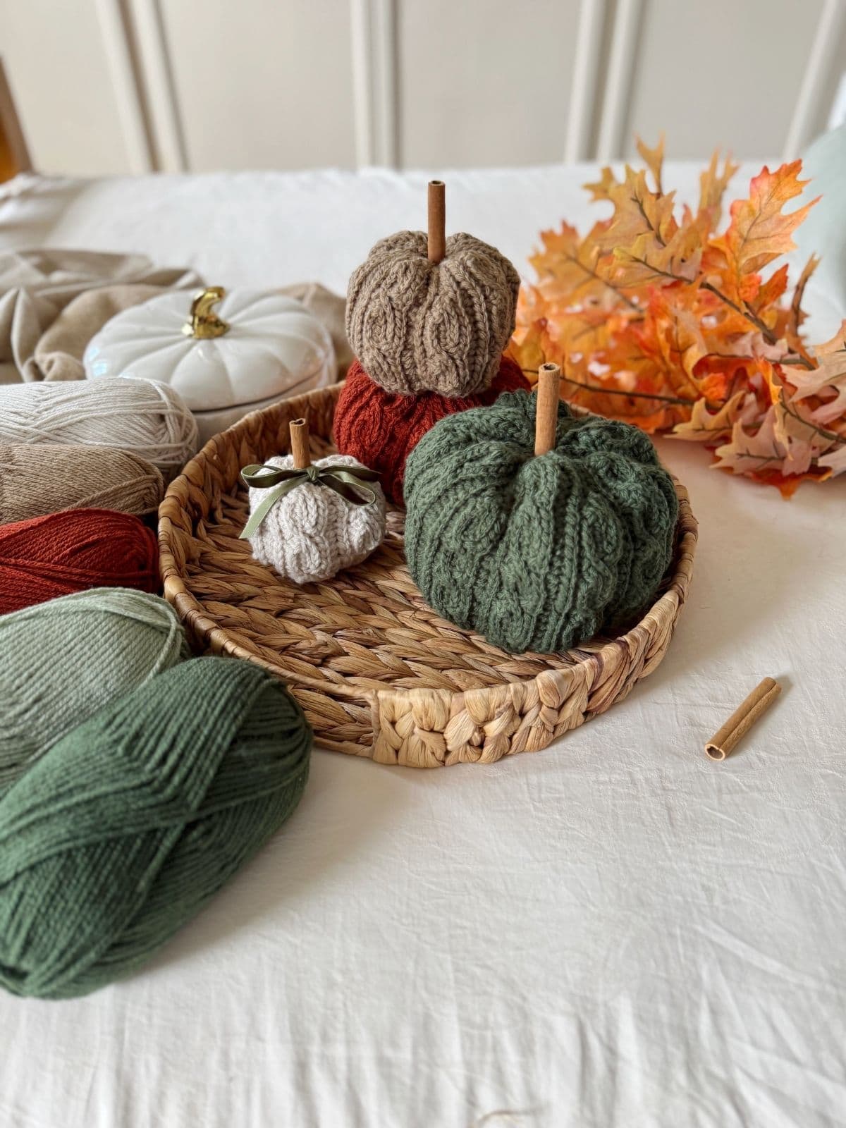 Three yarn pumpkins in a woven tray, surrounded by yarn balls, artificial autumn leaves, and a white pumpkin on soft fabric—perfect inspiration if you're searching for a crochet pumpkin pattern free to create your own cozy autumn décor.