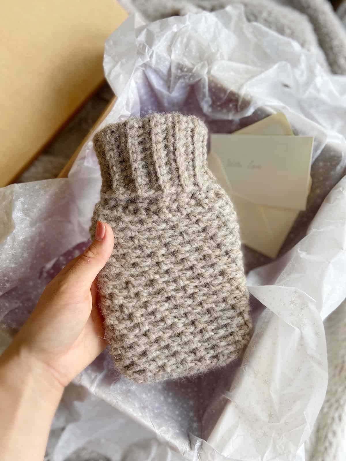 A hand holds a small, crocheted beige cover—perfect inspiration for fall crochet projects—over tissue paper in a box with envelopes nearby.