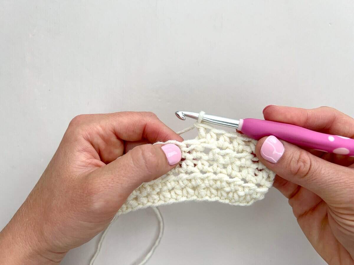 Two hands holding a white crochet piece, working front post and back post double crochet stitches with a pink-handled crochet hook against a plain white background.