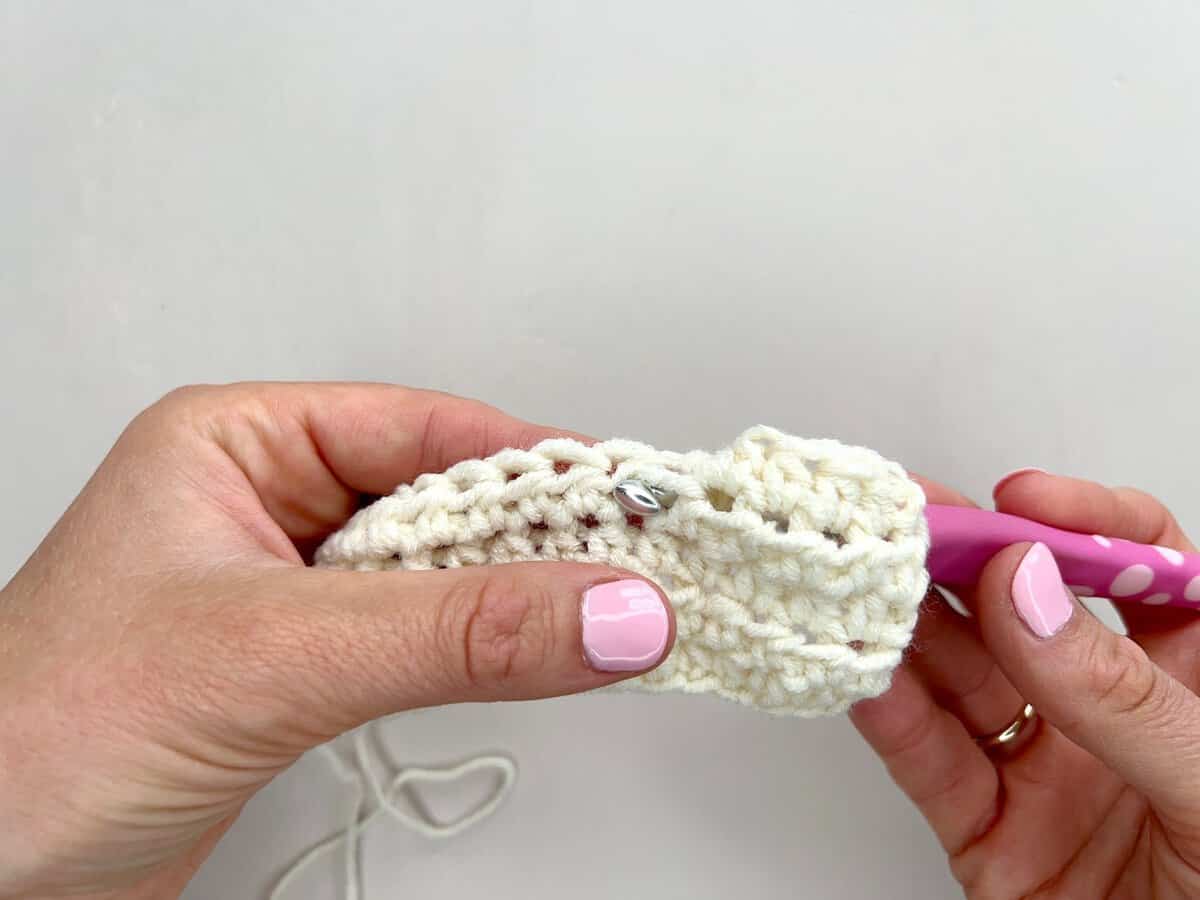 Two hands crocheting with white yarn and a pink crochet hook demonstrate front post and back post double crochet against a plain background.