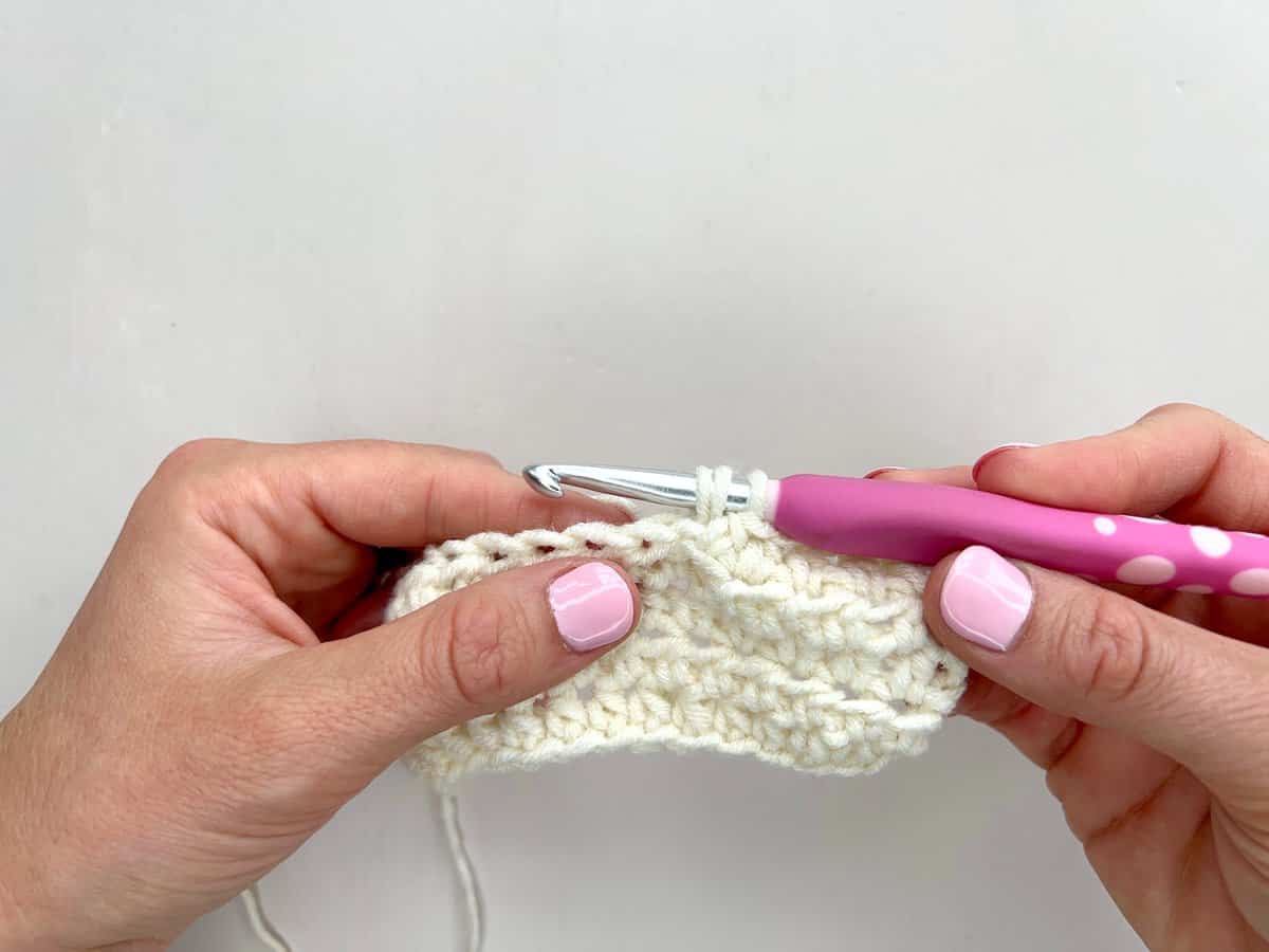 Two hands holding a crochet hook with a pink handle, demonstrating front post and back post double crochet techniques while working on a piece of white yarn against a plain background.