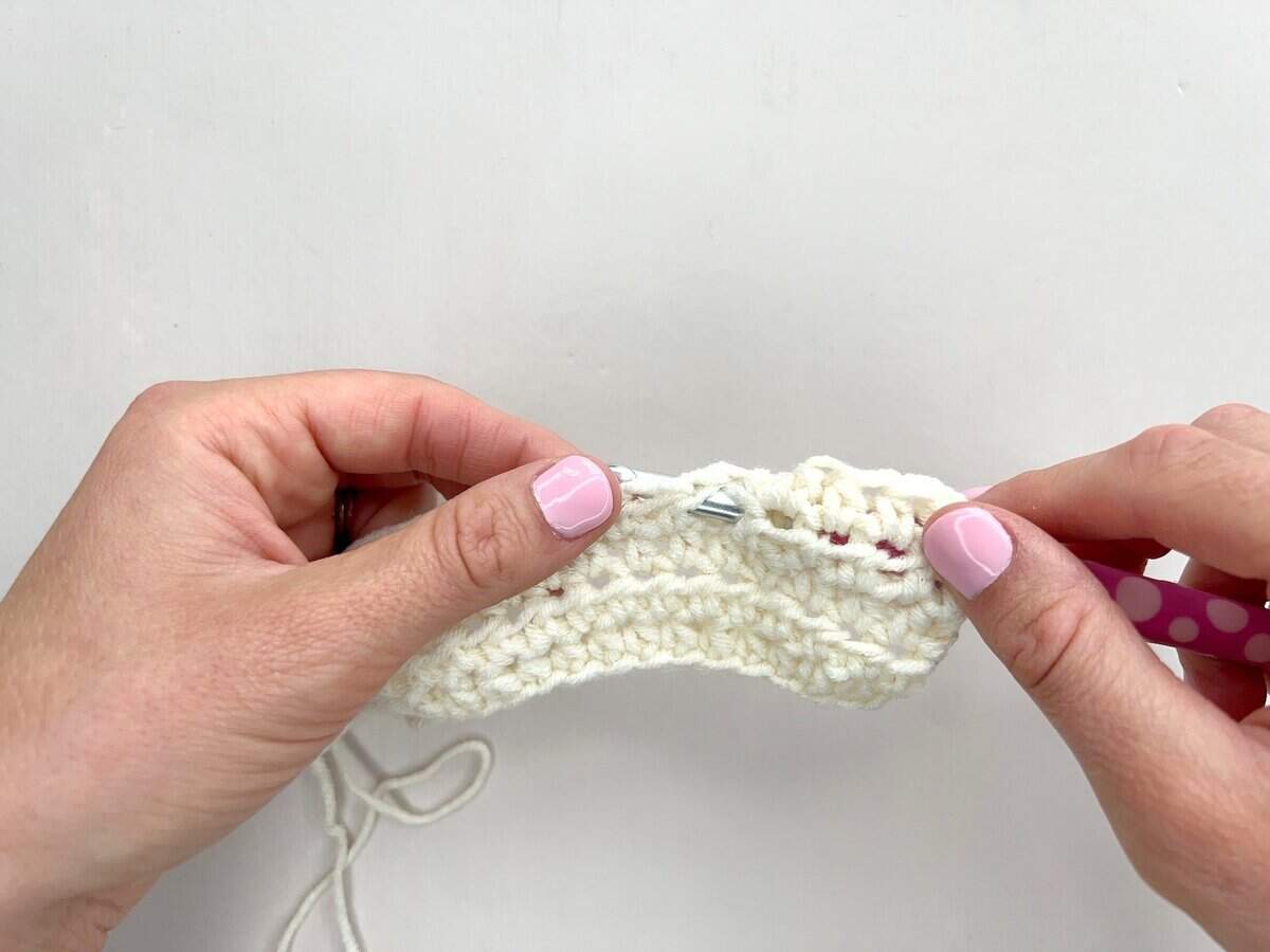 Two hands holding a crochet project in progress, demonstrating front post and back post double crochet stitches with a pink hook and white yarn against a plain white background.