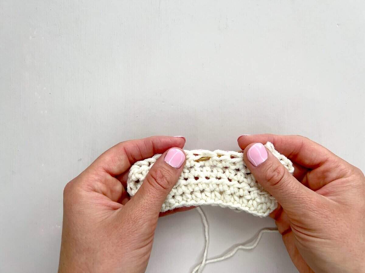 Two hands with light pink nail polish hold a piece of white crochet fabric, demonstrating front post and back post double crochet stitches with a yarn needle inserted into the work.