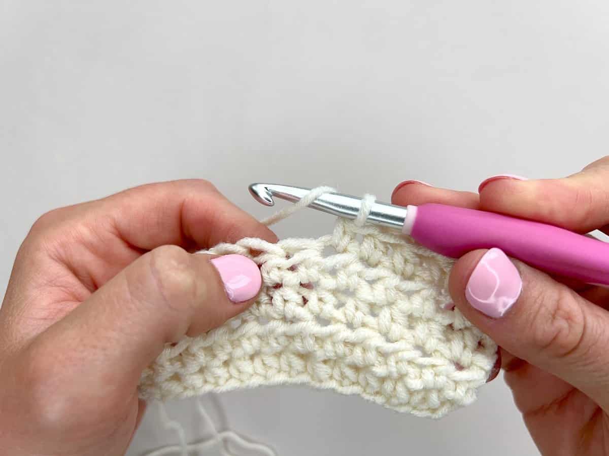 Close-up of hands crocheting with a pink-handled crochet hook and off-white yarn, demonstrating front post and back post double crochet stitches against a plain background.