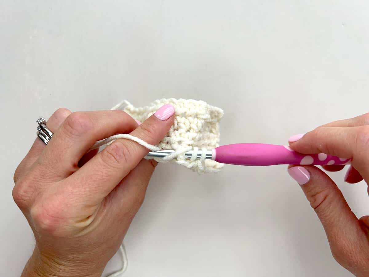 Two hands crocheting with white yarn and a pink crochet hook use front post and back post double crochet techniques against a white background.
