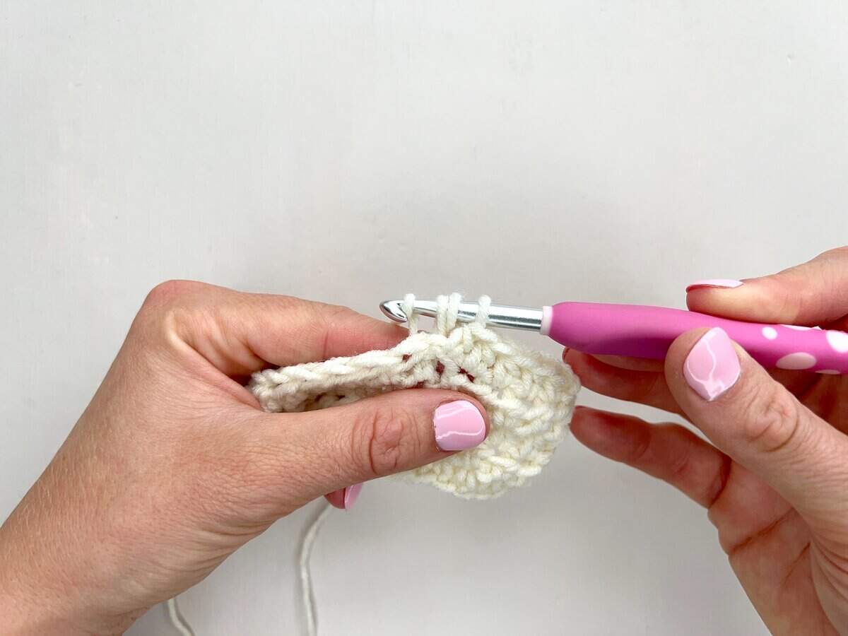 Two hands are crocheting with a pink and white crochet hook, skillfully working front post and back post double crochet stitches into a piece of off-white yarn against a plain background.