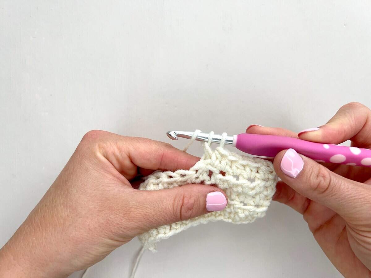 Two hands holding a white crocheted fabric, with the right hand using a crochet hook to create front post and back post double crochet stitches, forming neat rows of white yarn loops.