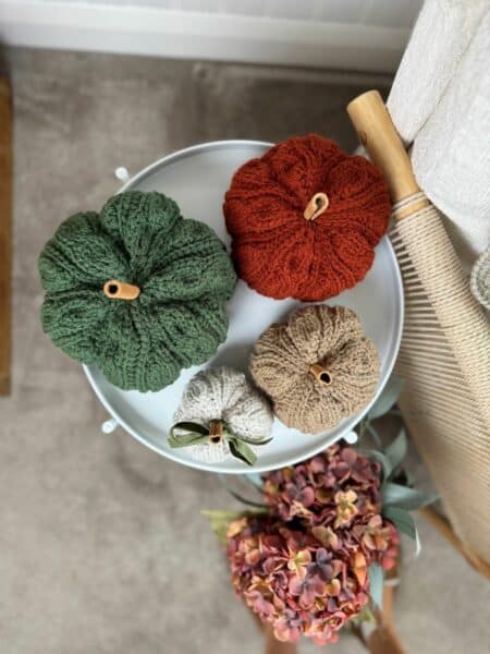 Four crochet pumpkins in green, orange, beige, and tan sit on a round white tray next to a bouquet of artificial hydrangeas and a wooden spindle—perfect inspiration if you’re searching for a crochet pumpkin pattern free online.