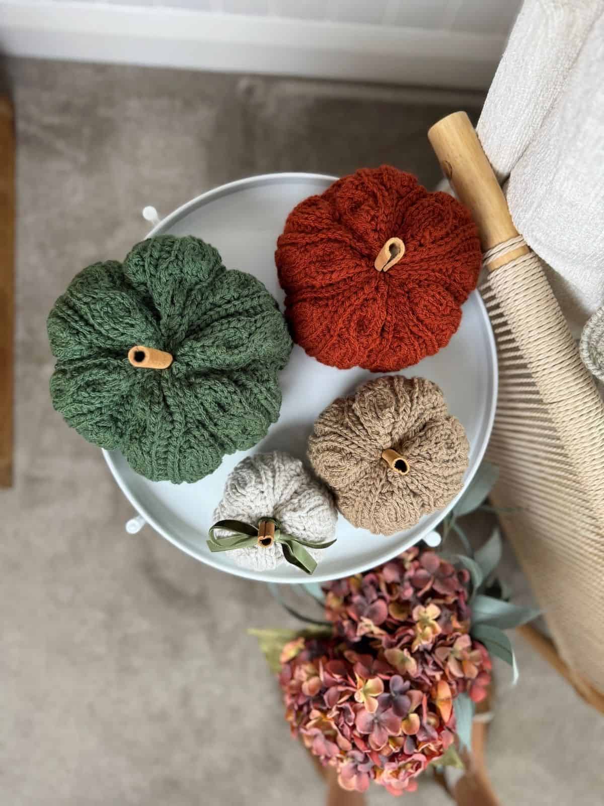 Four crochet pumpkins in green, orange, beige, and tan sit on a round white tray next to a bouquet of artificial hydrangeas and a wooden spindle—perfect inspiration if you’re searching for a crochet pumpkin pattern free online.