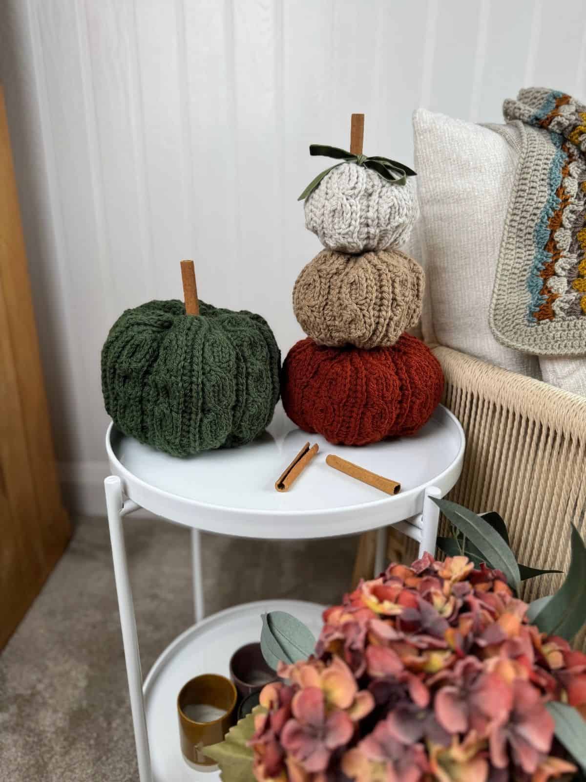 Three crocheted pumpkins—one green and two stacked in white, brown, and orange—sit on a round white table next to cinnamon sticks and a floral arrangement, inspiring cozy fall decor ideas with a crochet pumpkin pattern free to try.
