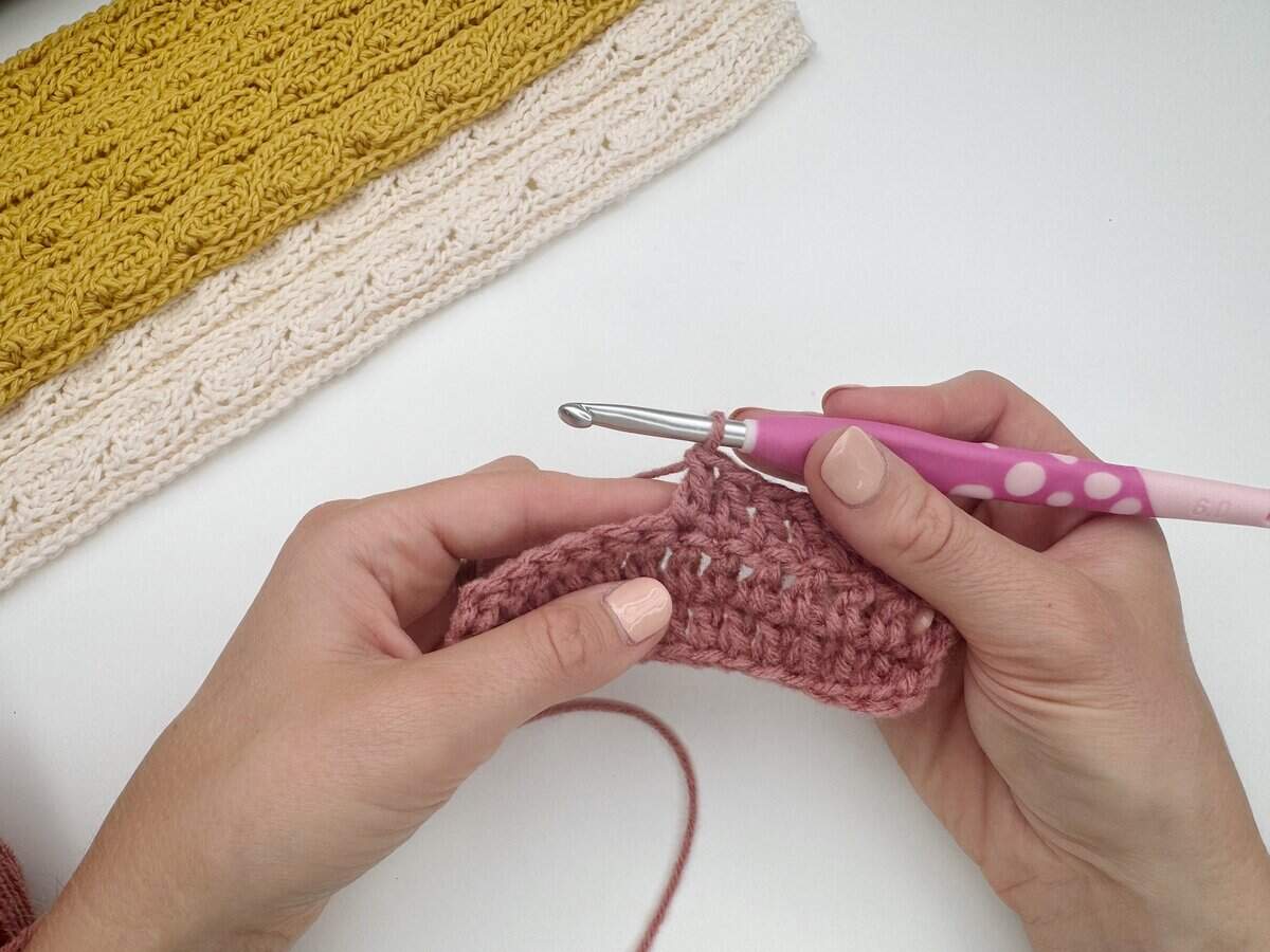 Hands holding a pink crochet hook, working with pink yarn, as completed yellow and cream crochet pieces rest on a white surface—perfect for learning how to do a double crochet stitch.