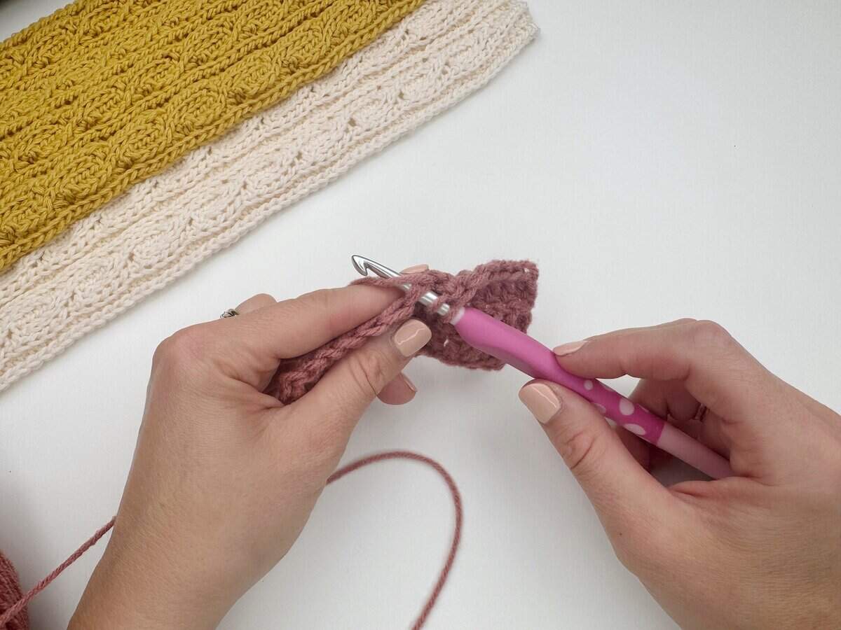 Hands crocheting pink yarn with a pink crochet hook, demonstrating how to do a double crochet stitch, with completed yellow and cream crochet pieces in the background on a white surface.