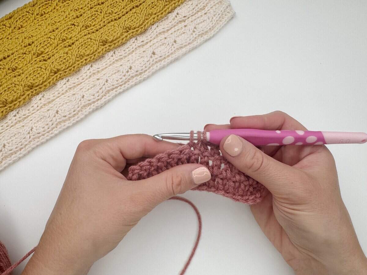 Two hands holding a crochet project with a pink and white crochet hook, demonstrating how to do a double crochet stitch with pink yarn. A finished yellow and cream crochet piece is above.