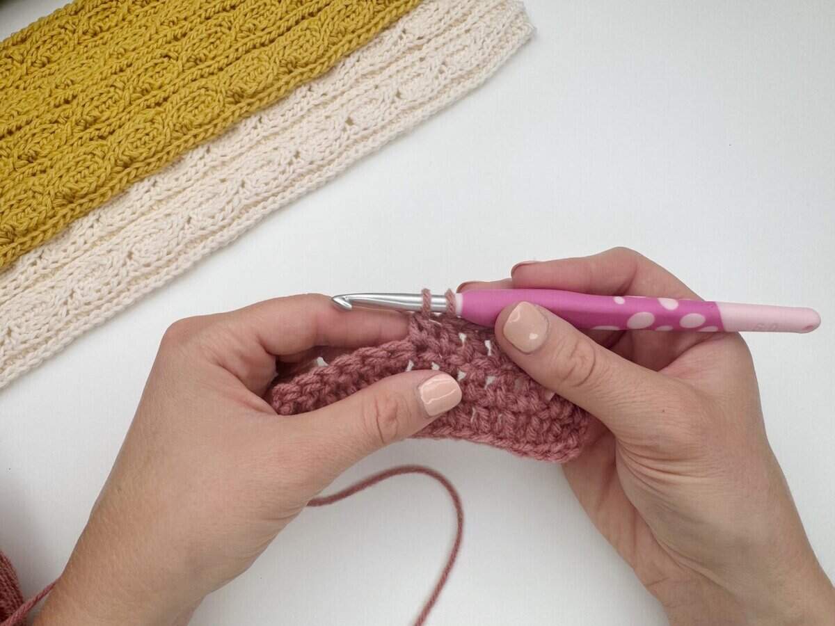 Close-up of hands showing how to do a double crochet stitch on a pink piece with a pink and white crochet hook; a yellow and cream crocheted piece is nearby on a white surface.