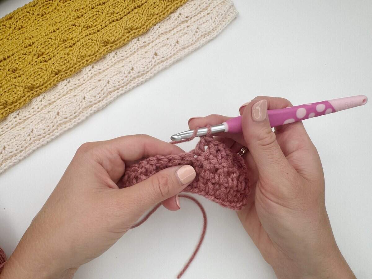 Close-up of hands crocheting with pink yarn and a pink crochet hook; a partially completed yellow and cream crochet piece lies on the table, perfect for learning how to do a double crochet stitch.