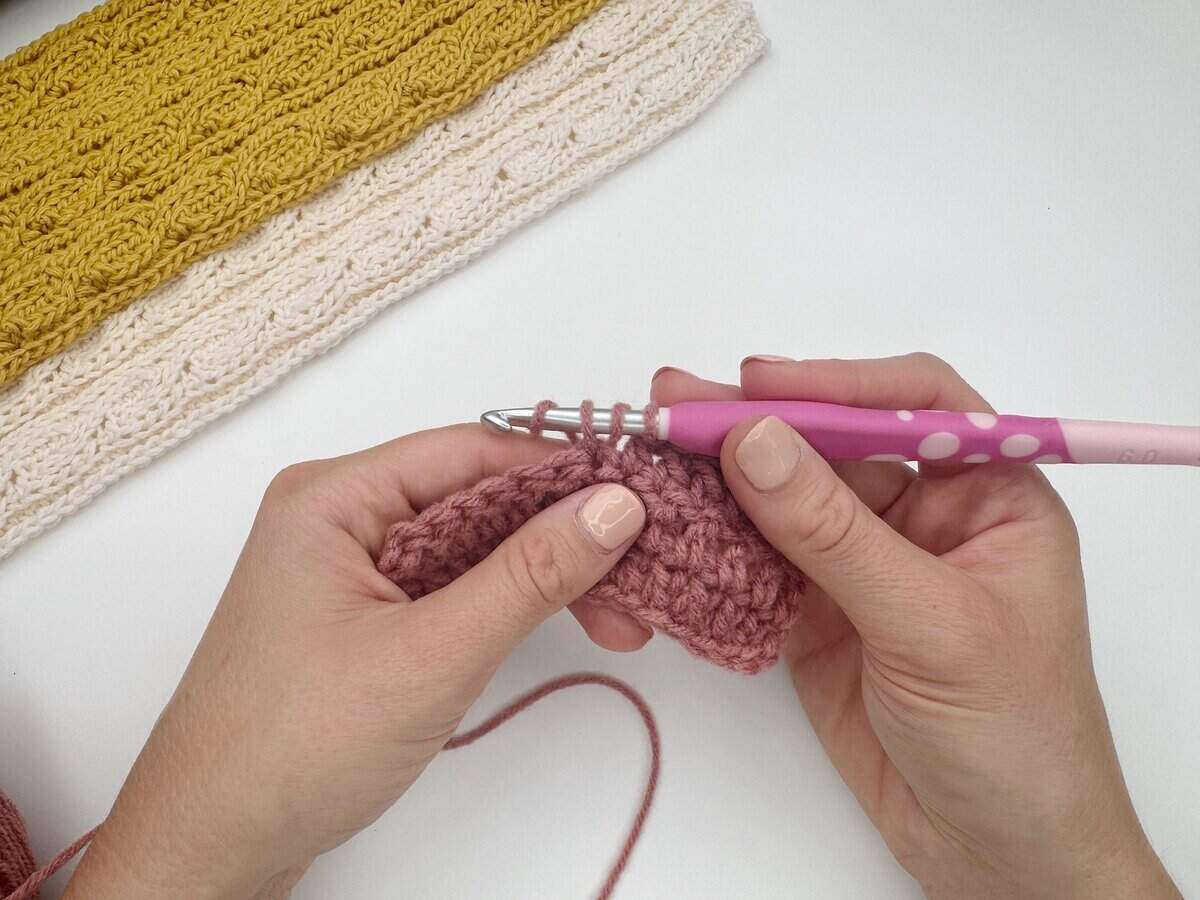 Two hands demonstrating how to do a double crochet stitch with pink yarn and a pink crochet hook, while finished yellow and cream crochet pieces rest in the background on a white surface.