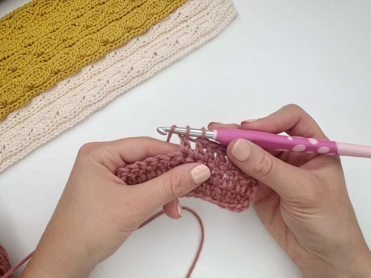 Close-up of hands demonstrating how to do a double crochet stitch with a pink-handled hook and pink yarn, as finished yellow and cream crochet pieces rest in the background on a white surface.