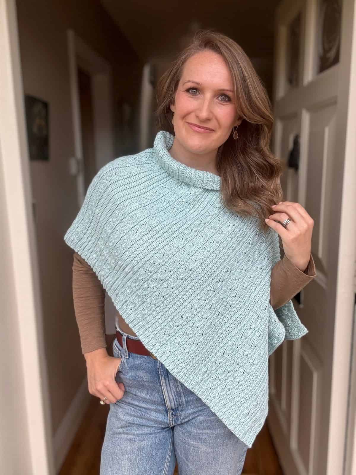 A woman stands indoors wearing a light blue poncho made with a free crochet poncho pattern, layered over a brown long-sleeve shirt and jeans, with one hand in her pocket and the other touching her hair.