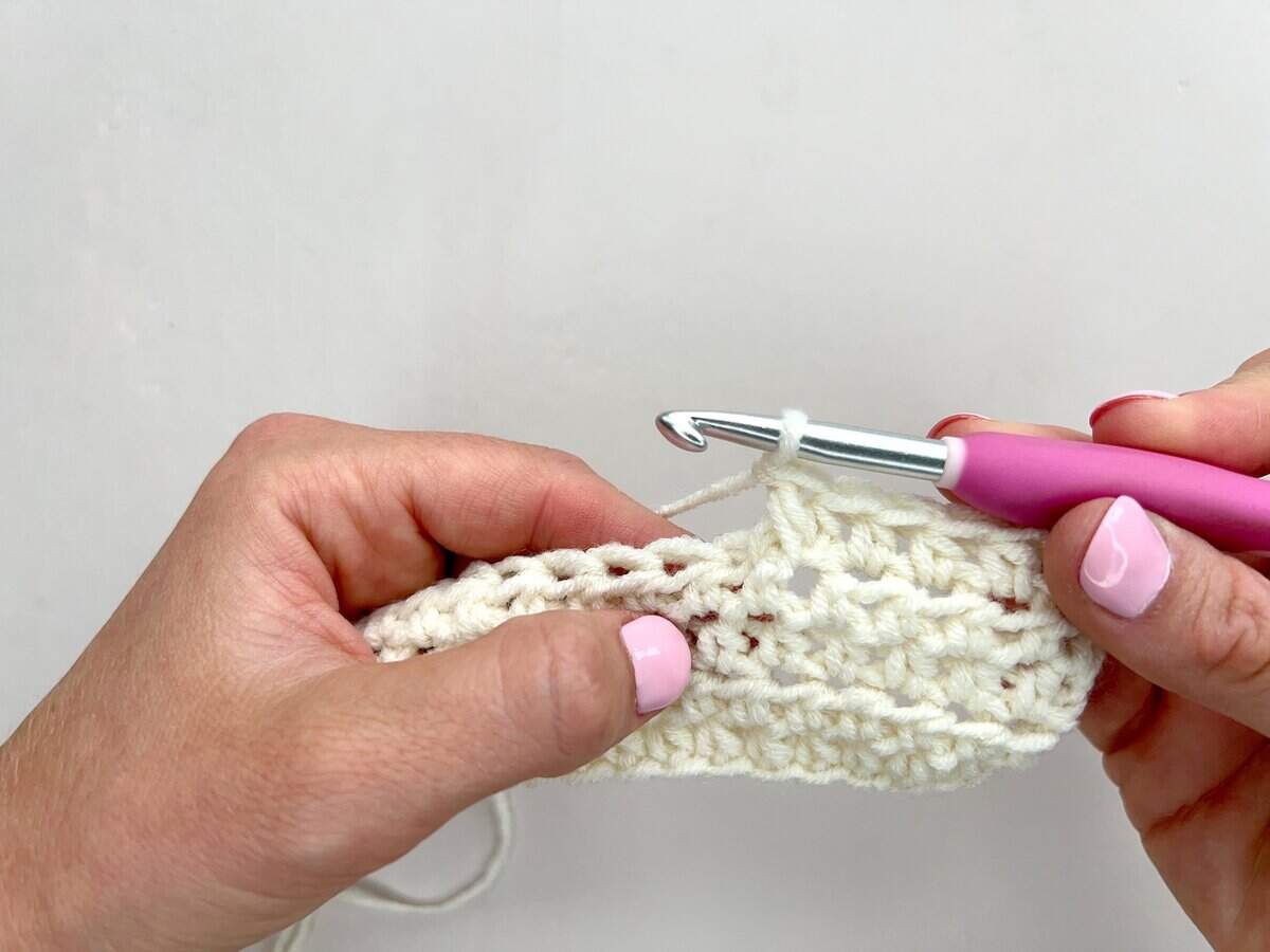 Two hands crochet with cream yarn using a pink-handled hook, creating a textured rectangular piece with front post and back post double crochet against a white background.