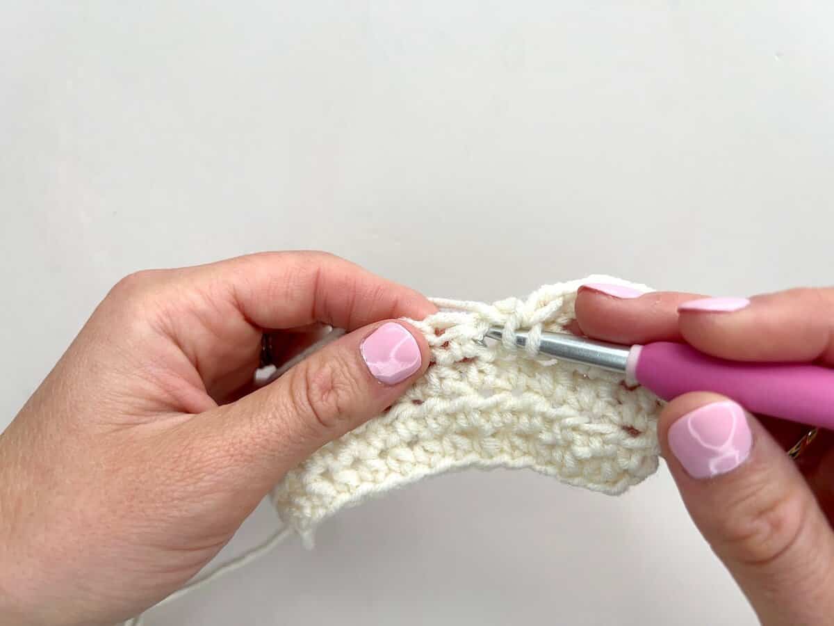 Close-up of hands crocheting with a pink-handled crochet hook and cream-colored yarn, skillfully working front post and back post double crochet stitches against a plain white background.