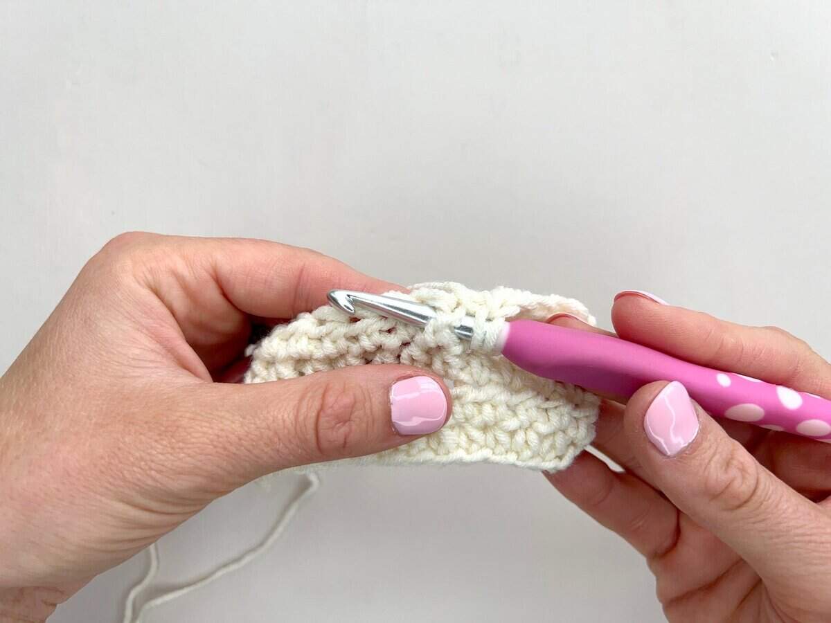 A person crochets with a pink-handled crochet hook and white yarn, working front post and back post double crochet stitches on a partially completed piece.