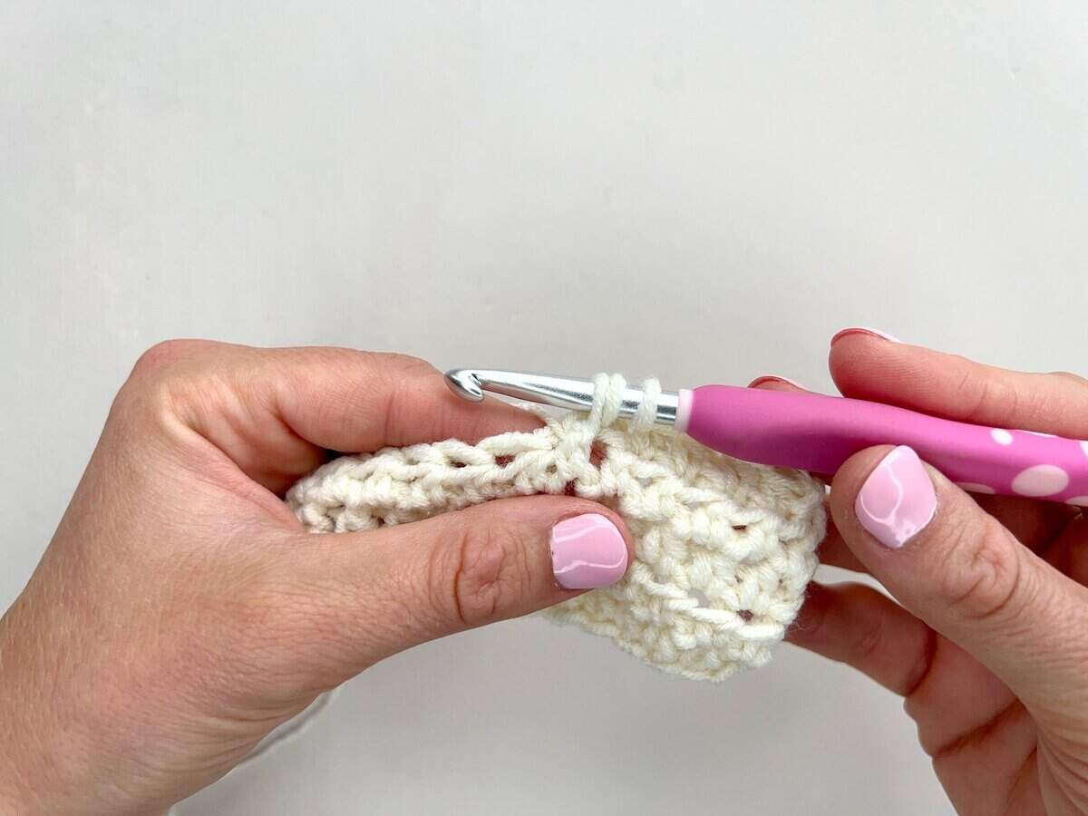 Close-up of hands crocheting front post and back post double crochet stitches with a pink-handled hook and white yarn against a plain background.
