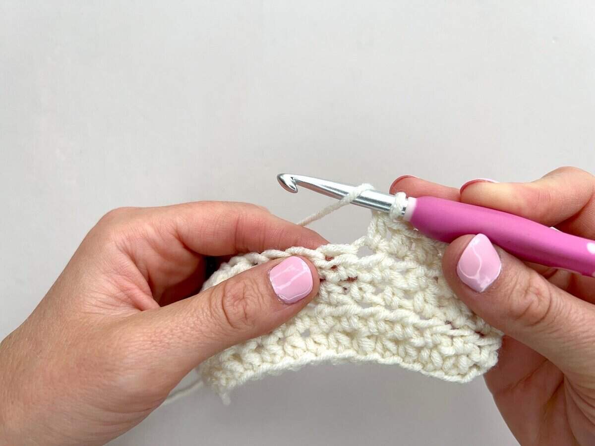 Close-up of hands skillfully crocheting front post and back post double crochet stitches with a pink-handled hook and cream-colored yarn against a plain background.