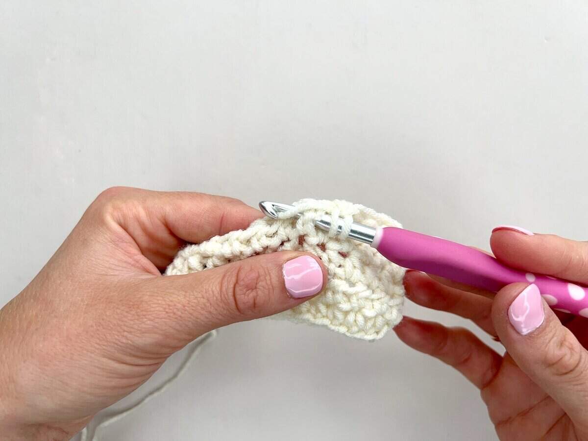 A person holding a crochet hook with a pink handle is working front post and back post double crochet stitches into a piece of white yarn.