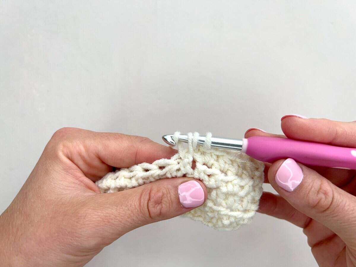 Close-up of hands crocheting with white yarn and a pink-handled crochet hook, skillfully working front post and back post double crochet stitches against a plain background.
