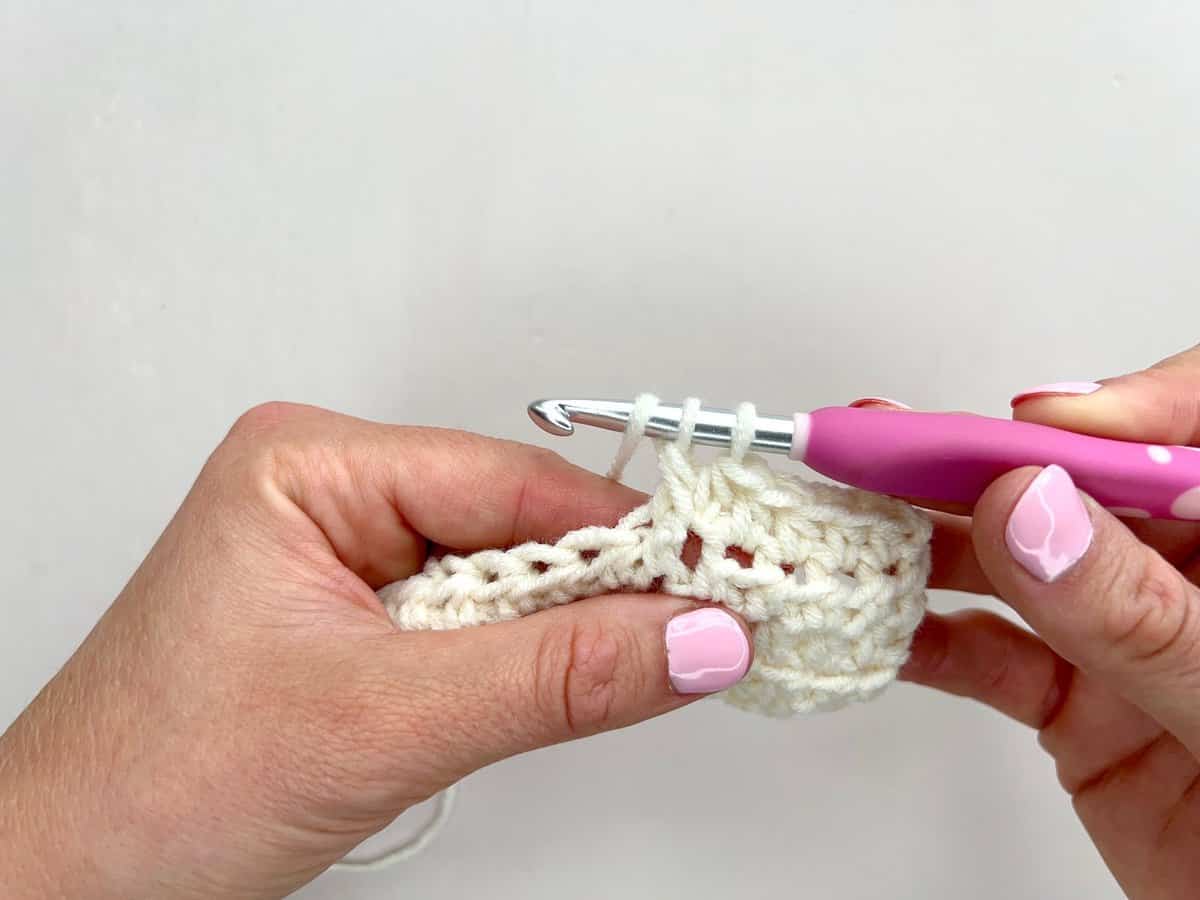 Close-up of hands crocheting with a pink-handled crochet hook and white yarn, forming front post and back post double crochet stitches against a plain background.