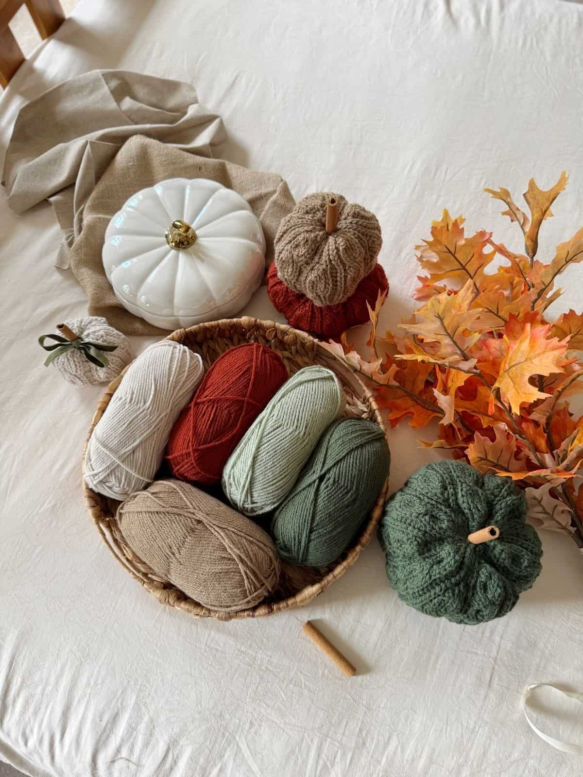 A basket of yarn balls in neutral and autumn colors sits on a bed with fabric pumpkins, artificial autumn leaves, and a white ceramic pumpkin—perfect inspiration for your next crochet pumpkin pattern free project.