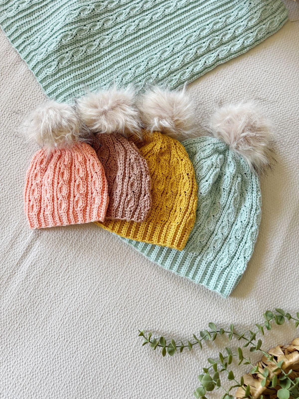 Four knitted hats in peach, beige, yellow, and light blue, each with a fluffy pom-pom, are arranged on a textured cream surface next to a matching scarf and a crochet fingerless mittens pattern free for download.