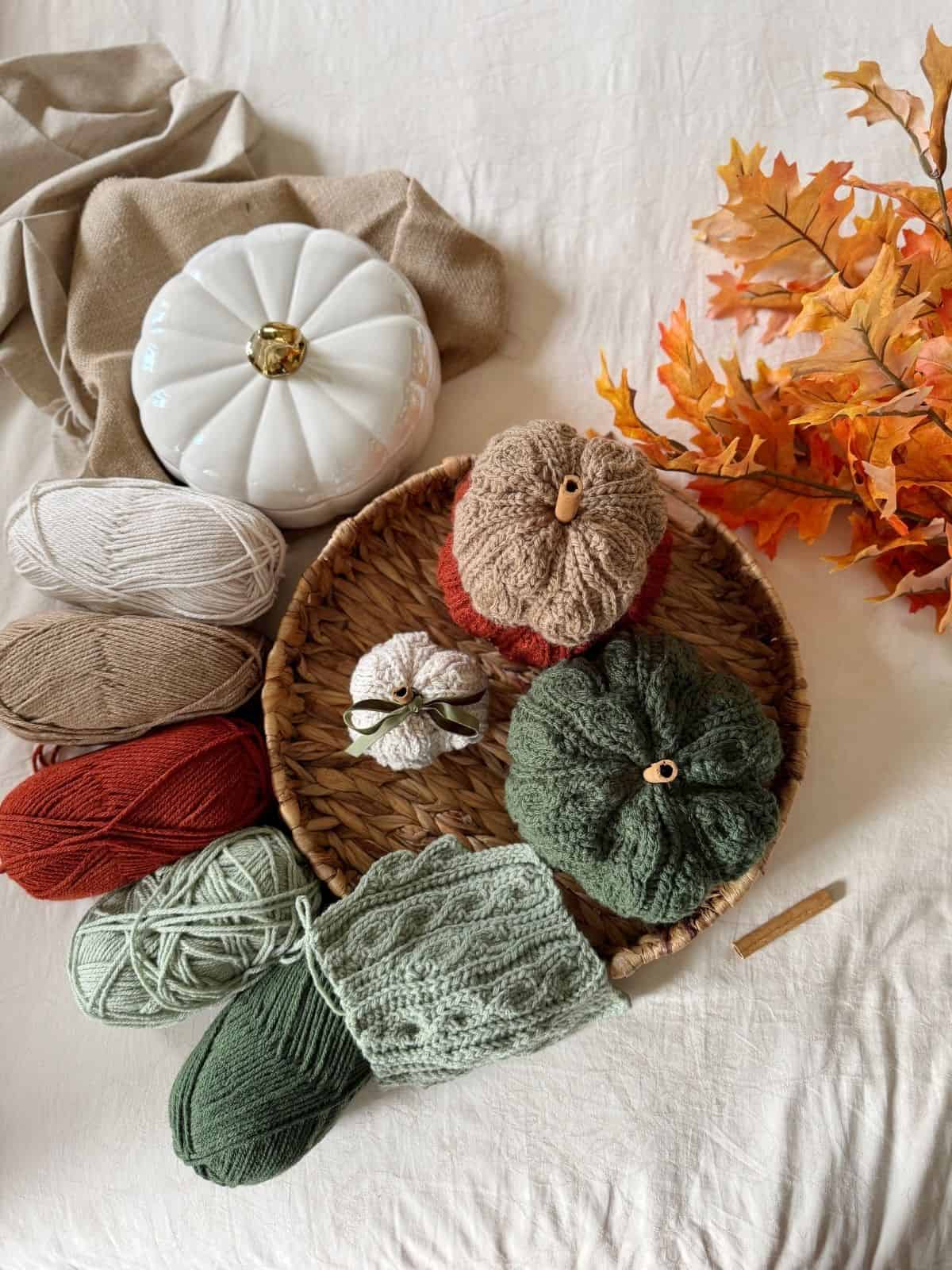 Yarn in various colors, crocheted pumpkins made using a crochet pumpkin pattern free, a ceramic pumpkin, and artificial autumn leaves arranged on a woven tray atop a fabric surface.