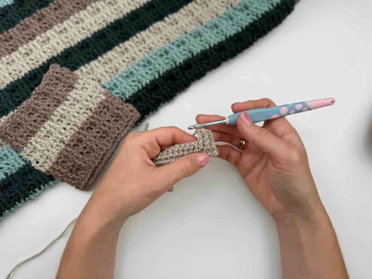 Close-up of hands crocheting with a light blue and pink hook, working on a beige piece next to a striped crocheted fabric in shades of beige, blue, and dark gray using alternating front and back post double crochet stitches.