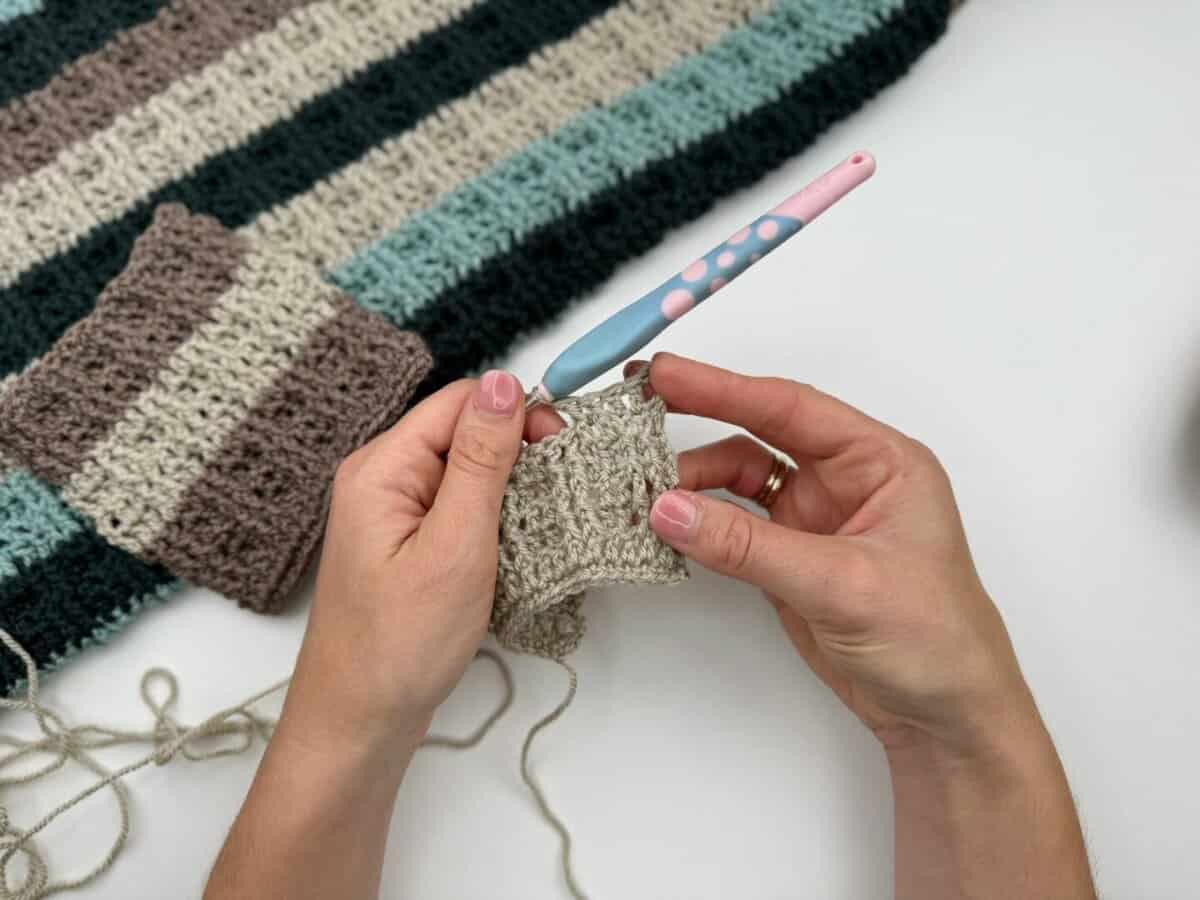 Hands are crocheting a piece of beige yarn with a pink and blue crochet hook, using alternating front and back post double crochet, next to a striped crochet blanket in brown, green, and beige tones.
