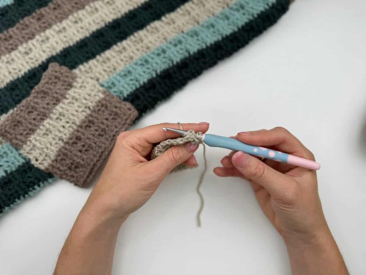 Hands holding a crochet hook and working with beige yarn, creating a striped crochet fabric using alternating front and back post double crochet, with a crochet swatch in the background.
