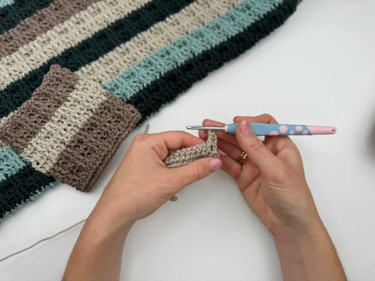 Hands holding a crochet hook and yarn, working on a beige piece using alternating front and back post double crochet, next to a striped, crocheted fabric in brown, blue, and cream tones on a white surface.