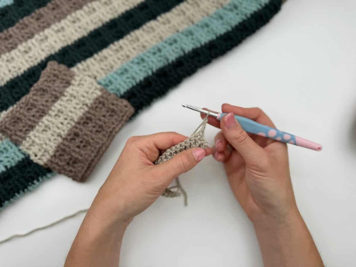 A person crocheting with a pink and blue crochet hook, working on a beige piece using alternating front and back post double crochet next to a striped crocheted fabric on a white surface.