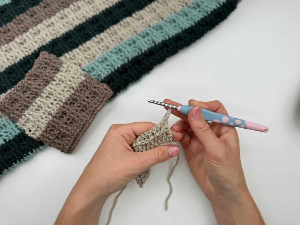 Hands crocheting a piece of beige yarn with a pink and blue crochet hook, demonstrating alternating front and back post double crochet; a striped crocheted fabric is visible in the background.