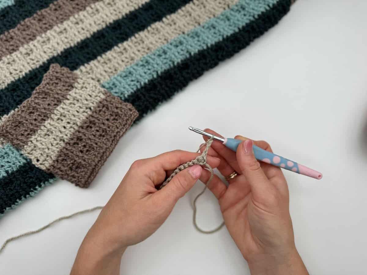 Hands holding a crochet hook and working with light-colored yarn next to a striped crocheted fabric in shades of brown, blue, and black featuring alternating front and back post double crochet stitches.