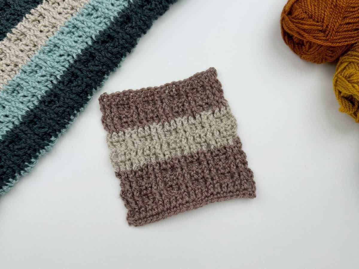 A small crocheted swatch, made using alternating front and back post double crochet with two brown stripes and one beige stripe, lies on a white surface next to a striped crochet piece and two balls of yarn.