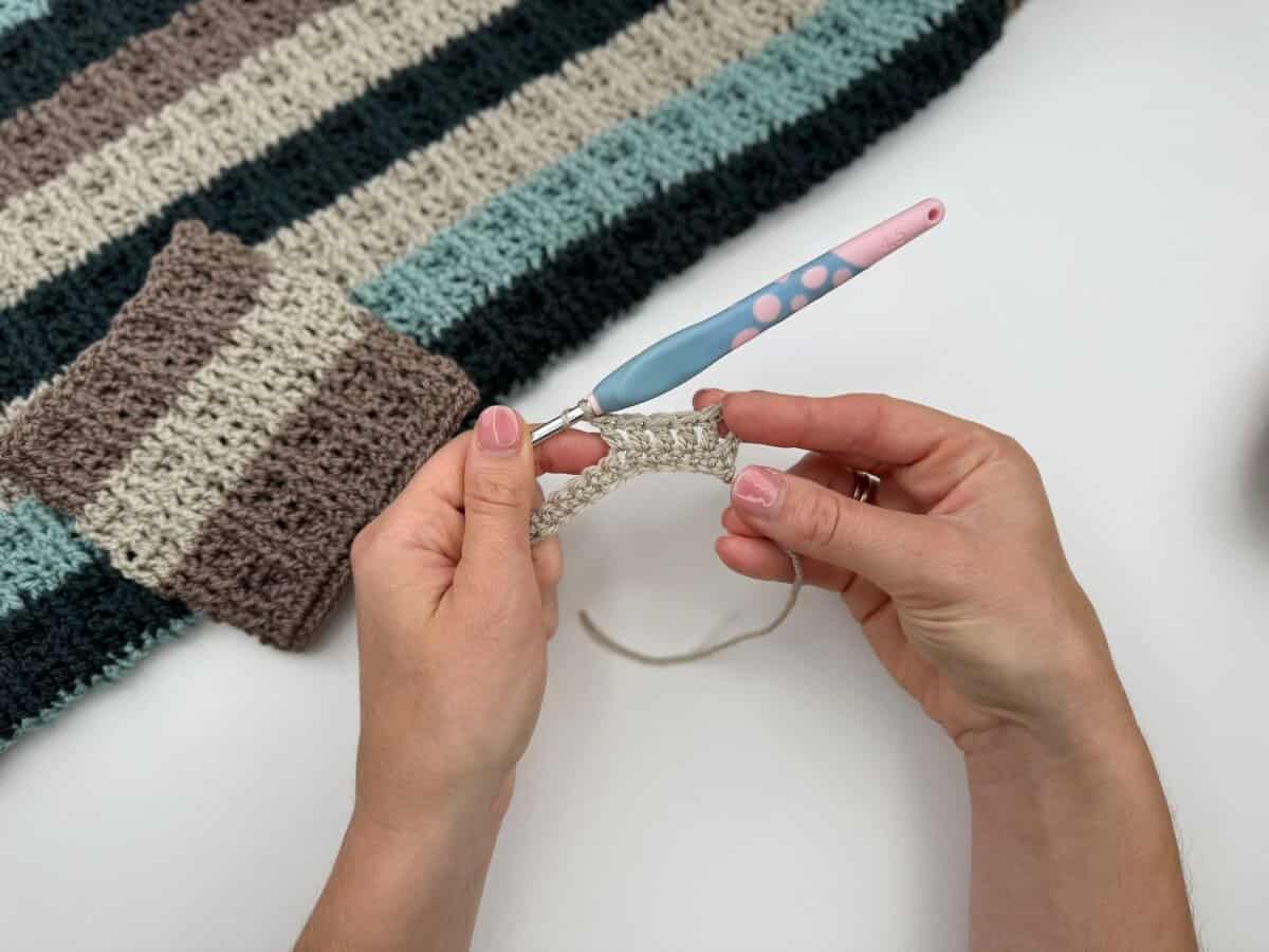 Hands holding a crochet hook and working beige yarn, demonstrating alternating front and back post double crochet; a striped crochet project in brown, blue, and cream rests on the table in the background.