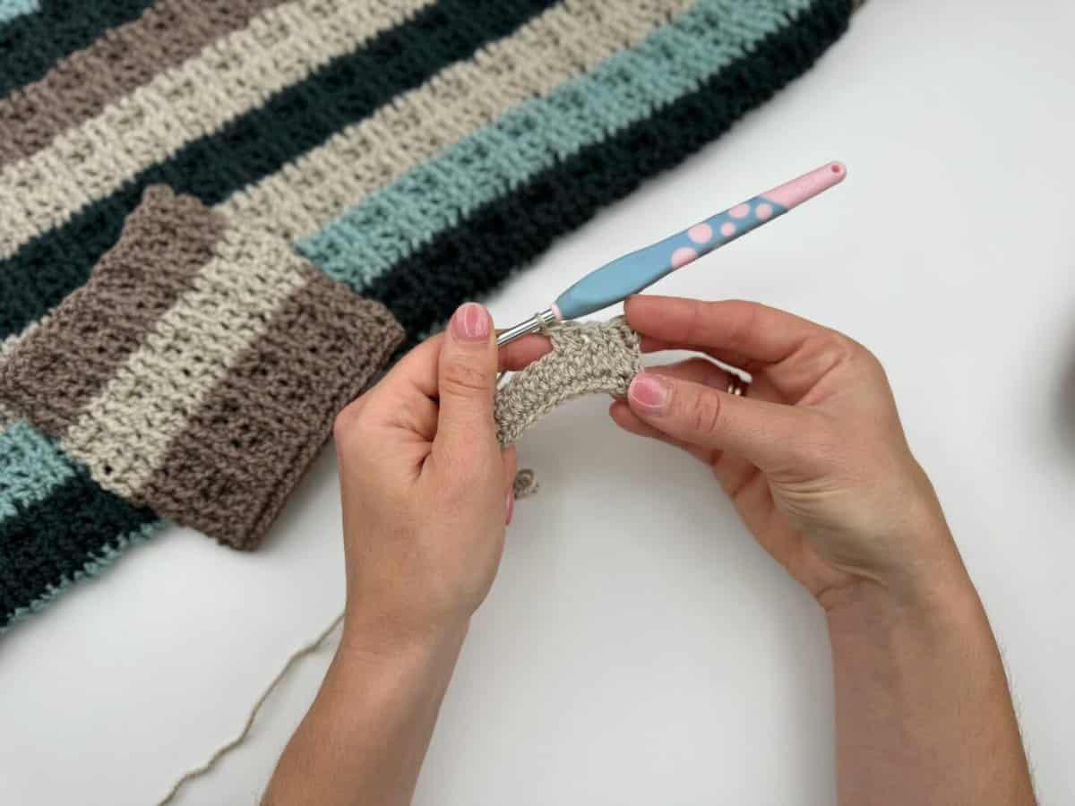 Two hands crochet with a blue and pink hook, working beige yarn in alternating front and back post double crochet. Nearby are a striped crocheted fabric piece and a small finished square in similar colors.