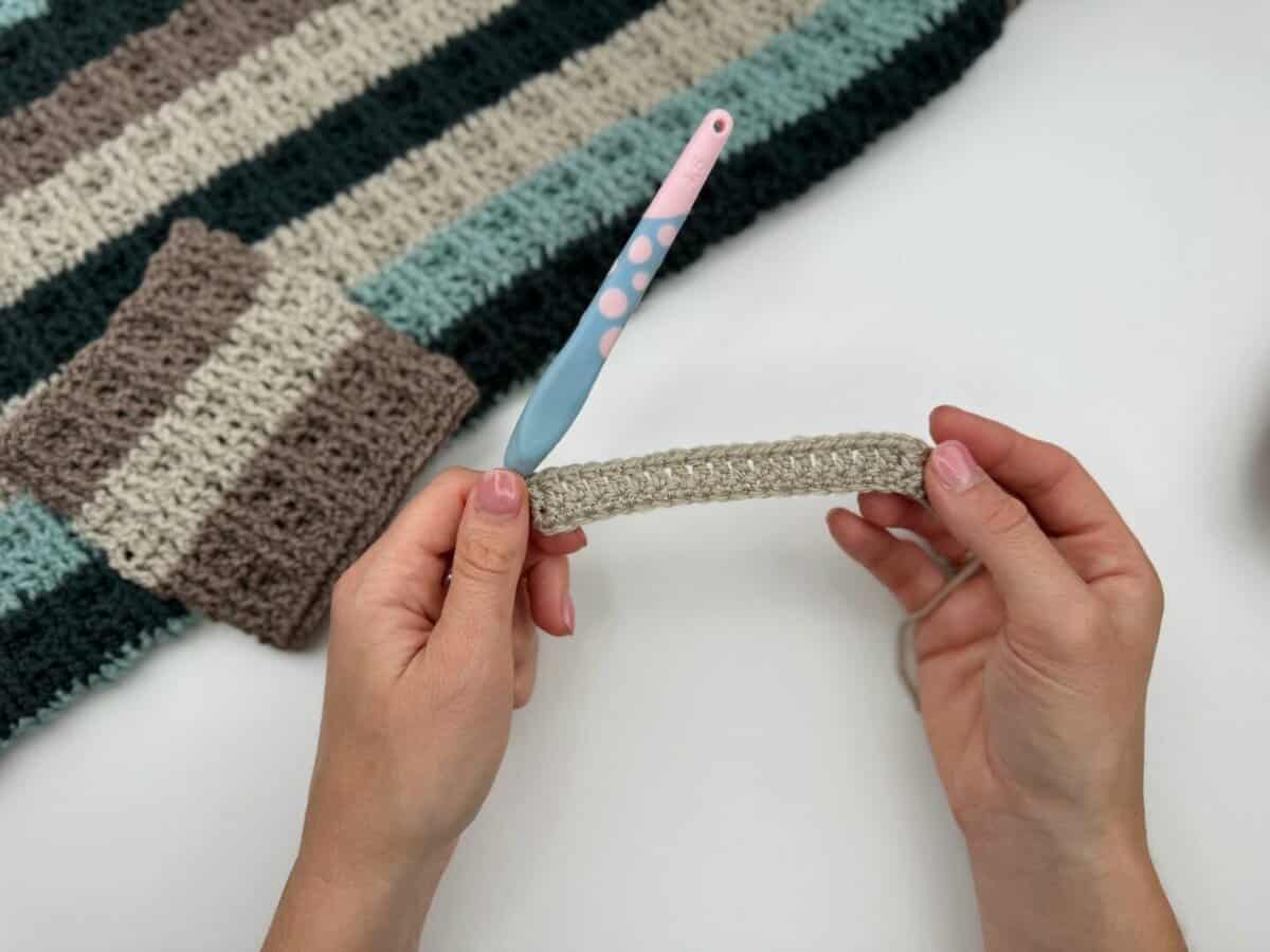 A person holds a small crocheted strip with both hands; a crochet hook and a larger striped crochet piece, featuring alternating front and back post double crochet, are on the table.